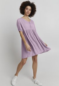 Hellblaues, kurzärmeliges Kleid mit V-Ausschnitt und Knopf-Detail. Flüssiger, geraffter Rock aus strukturiertem Stoff. Getragen mit weißen Sneakers.