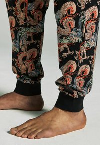 Schwarze Hose mit einem bunten Drachenmuster, ausgestattet mit elastischen Bündchen und einer weichen Textur. Die nackten Füße des Models sind unten sichtbar.
