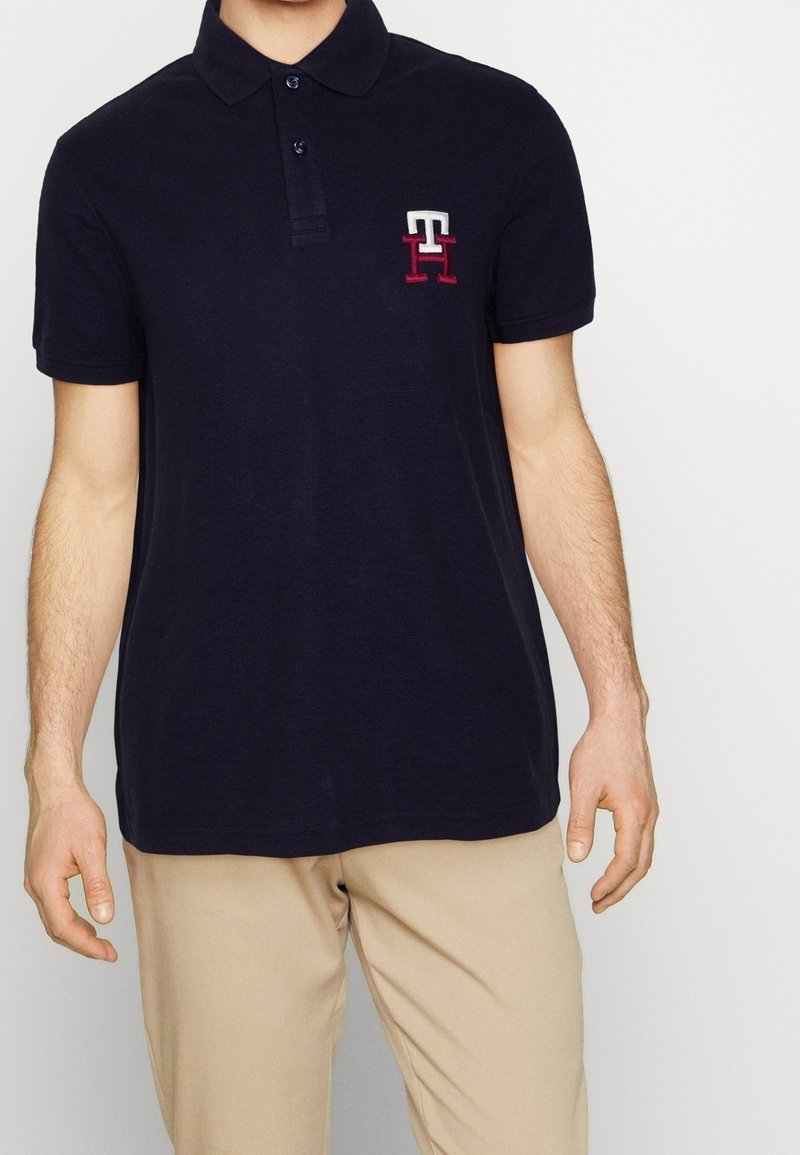 Polo shirt navy en coton avec un col, une patte de boutonnage à deux boutons et un logo brodé en blanc et bordeaux sur la poitrine gauche.