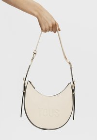 Bolso de piel beige con forma de media luna, ribete negro, logo "TOUS" grabado en la parte frontal y una correa ajustable.