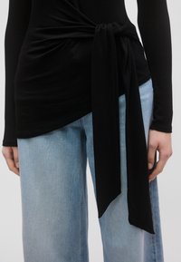 Bluza neagră cu mâneci lungi și detaliu cu șnur în zona taliei, asortată cu blugi largi de culoare albastru deschis. Materialul este neted și oferă o ușoară elasticitate.