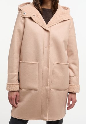 Femme portant un manteau à capuche rose pâle avec de grandes poches avant et des manches retroussées sur un pull foncé.