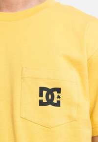 Žlté bavlnené tričko s okrúhlym výstrihom, s čiernym logom DC vytlačeným na ľavej náprsnej vrecku. Mäkká textúra, ležérny dizajn.