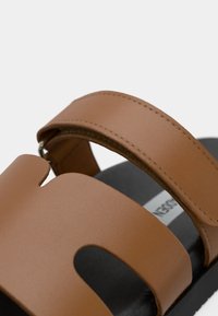 Sandale synthétique marron avec un design à découpes, une large bride et une semelle noire texturée. Couture apparente le long des bords de la bride.