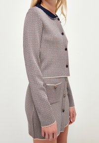 Kvinna iklädd en matchande set av kappa och kjol med houndstooth-mönster, mörka knappar och kontrasterande detaljer, stående mot en enkel bakgrund.