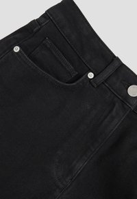 Close-up of black denim jeans front pocket with belt loop, metal rivets, and stitching details on plain background.