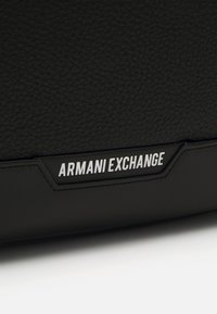 Zwarte gestructureerde leren tas met een opvallend wit "Armani Exchange" logo op een rechthoekig label, met strakke lijnen en een minimalistisch ontwerp.