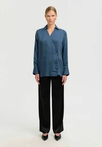 Camisa azul de satén con cuello en pico, mangas largas y un botón decorativo, combinada con pantalones negros de cintura alta y tacones puntiagudos.