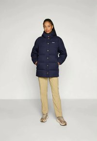 Casaco isolado azul-marinho com gola alta, fecho frontal por botões e dois bolsos laterais. Usado com calças bege e ténis de cor clara.