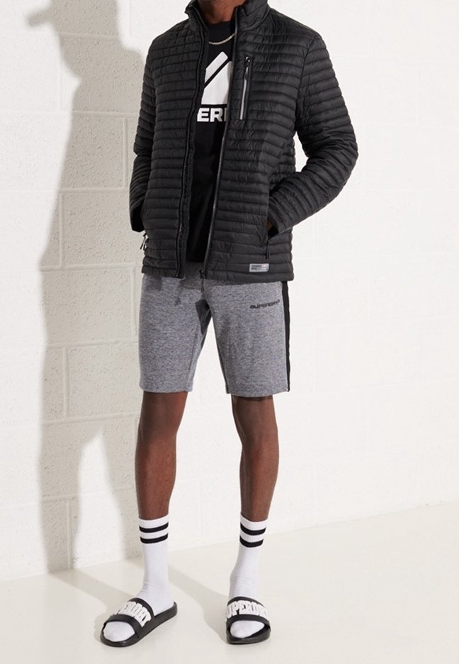 Veste matelassée noire avec poche zippée, t-shirt graphique noir, shorts gris avec bande latérale, chaussettes blanches avec rayures noires et sandales noires à enfiler.