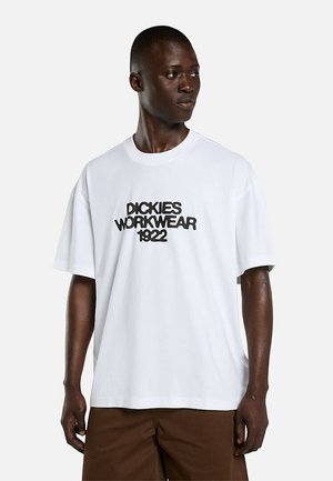 Hombre con camiseta blanca extragrande con texto negro "Dickies Workwear 1922", combinado con pantalones marrones, mirando hacia un lado.