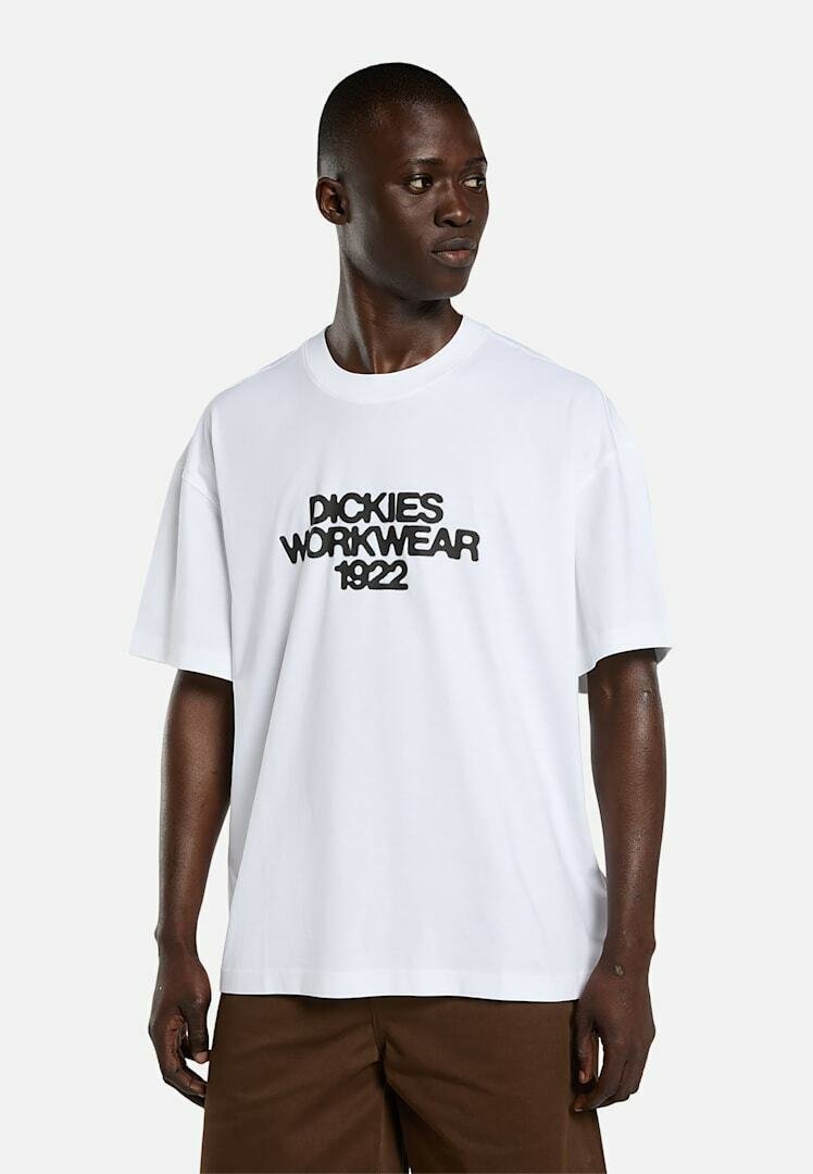 Mann trägt weißes übergroßes T-Shirt mit schwarzem Schriftzug „Dickies Workwear 1922“, kombiniert mit brauner Hose, schaut zur Seite.