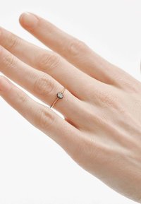 Anillo delgado de oro con una pequeña gema redonda y clara en el centro. La mano muestra una textura de piel natural con esmalte de uñas suave.