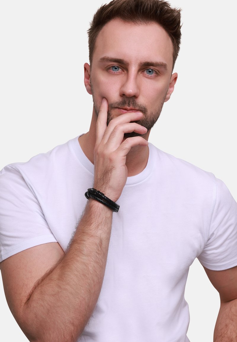 Black braided leather bracelet on wrist, worn with a plain white t-shirt. Straight-cut collar and solid color with smooth texture.