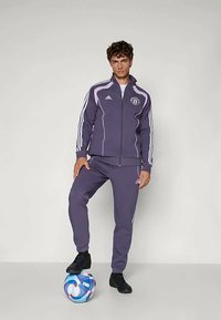 Lila Adidas-träningsoverall med vita ränder, som har en logotyp på vänster bröst. Matchad med svarta skor och en flerfärgad fotboll.