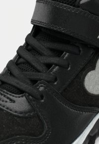 Nærbilde av svarte sneakers med tykke snorer, mesh-stofftunge og en reflekterende hjerteformet detalj på siden.