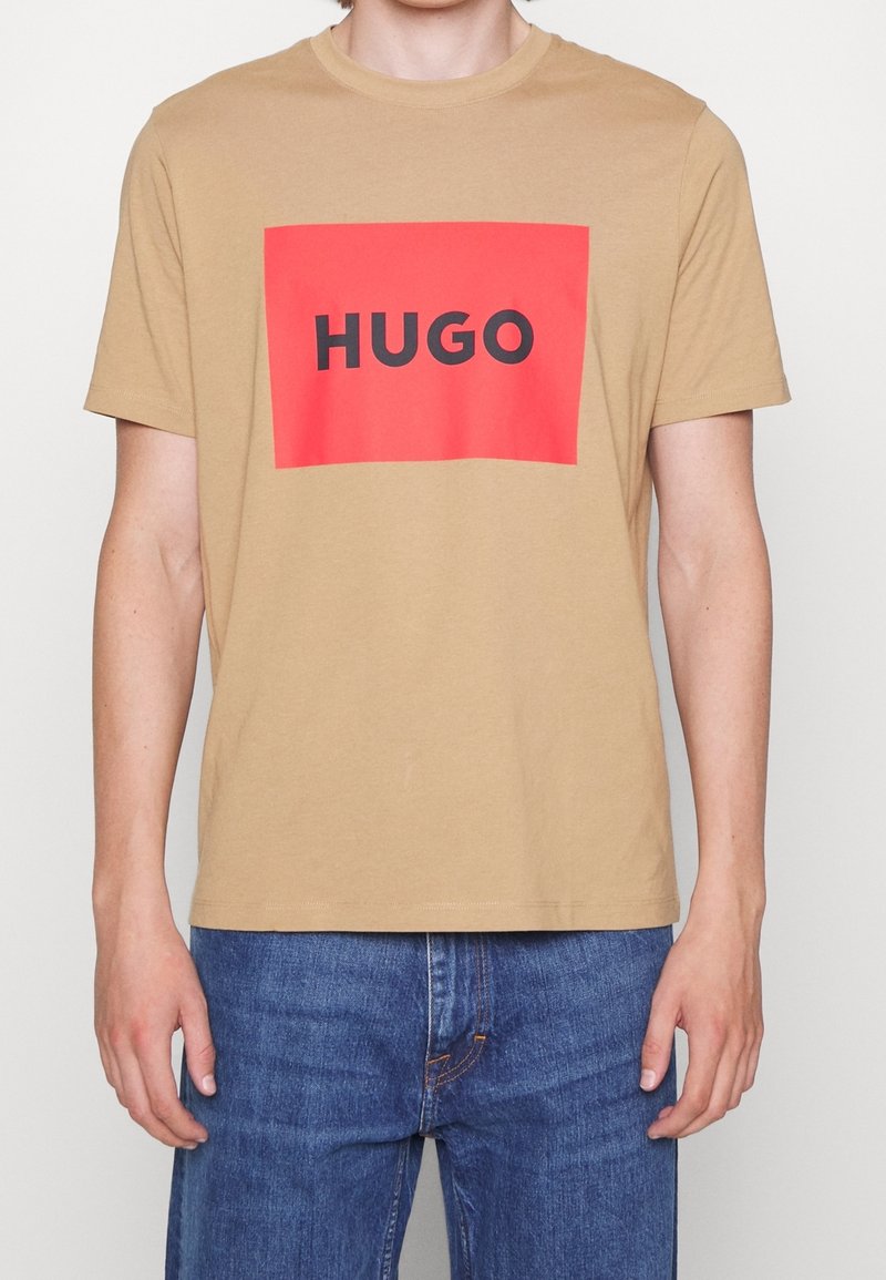 T-shirt en coton beige avec un rectangle rouge audacieux portant le mot "HUGO" en lettres noires. Manches courtes et design classique à col rond.