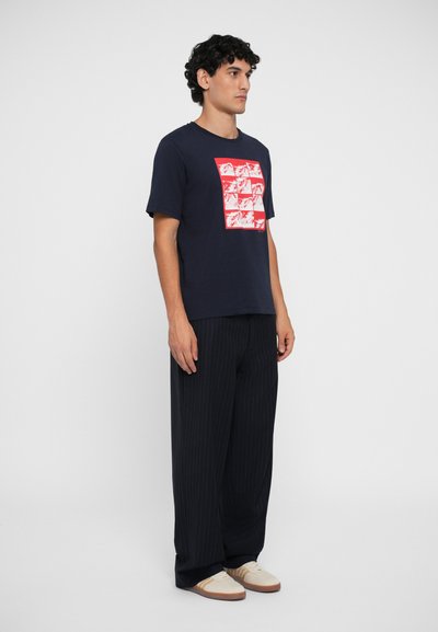 Camiseta de algodón azul marino con un estampado gráfico rojo, combinada con pantalones anchos de rayas negras. El modelo está de lado, vistiendo zapatos color beige.
