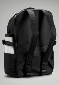 Svart ryggsäck gjord av slitstark nylon, med justerbara remmar, en sidoficka som rymmer en vit och svart vattenflaska och en dubbel dragkedja.