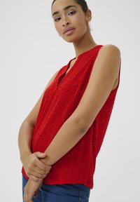 Rote ärmellose Bluse mit V-Ausschnitt, die geometrische Ausschnittmuster und einen strukturierten Stoff aufweist. Kombiniert mit blauen Jeans.