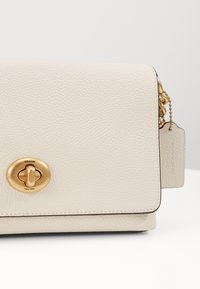 Sac à main en cuir texturé beige avec une fermeture à rabat, des accessoires en ton doré et une étiquette amovible. Présente un logo signature sur le fermoir.