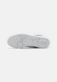 Semelle extérieure de sneaker blanche en caoutchouc texturé pour une adhérence, dotée d'une rainure de flexion circulaire et d'un détail de marque sur le côté.