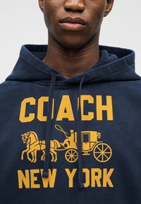 Dunkelblauer Hoodie mit gelbem Text "COACH NEW YORK" und einer Grafik einer Pferdekutsche mit einem Fahrer, der eine Peitsche hält.