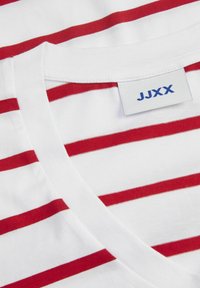 Camiseta blanca con rayas rojas horizontales, diseño de cuello en V, material de algodón y una etiqueta con "JJXX" impresa en azul.