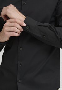 Schwarzes Langarmshirt mit Knopfleiste, glatter Textur, tailliertem Design und Knopfmanschetten. Verfügt über passende schwarze Knöpfe aus dem gleichen Stoff.
