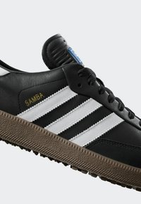 Czarne skórzane sneakersy z trzema białymi paskami, teksturowaną brązową gumową podeszwą, wyprofilowanym czarnym językiem oraz napisem "SAMBA" w złotym kolorze po boku.