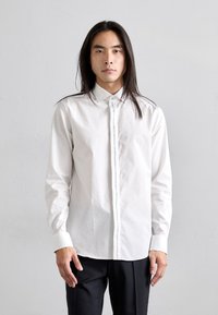 Camicia bianca a maniche lunghe in cotone con colletto button-down, caratterizzata da una patta frontale e sottili accenti blu lungo la chiusura.