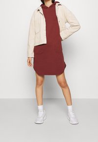 Beżowa kurtka z sztruksu noszona na burgundowej sukience z kapturem z dwoma przednimi kieszeniami. Białe sneakersy i skarpetki do kostek dopełniają całość.