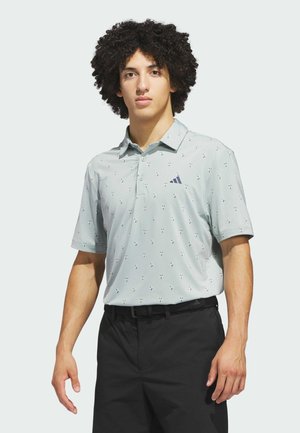 Junger Mann mit lockigem Haar, der ein hellgraues, gemustertes Poloshirt und schwarze Hose trägt und vor einem schlichten Hintergrund steht.