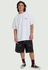 Camiseta de algodón blanca de gran tamaño con un pequeño logo negro, combinada con pantalones cortos negros con costuras blancas y bolsillos laterales, y zapatillas negras.