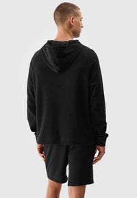 Schwarze Kapuzenjacke mit langen Ärmeln, lockere Passform und weichem Material. Kombiniert mit passenden schwarzen Shorts, von hinten gesehen.