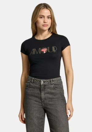 Donna che indossa una t-shirt aderente nera con la scritta "AMOUR" e una grafica di labbra rosse, abbinata a jeans grigi a vita alta.