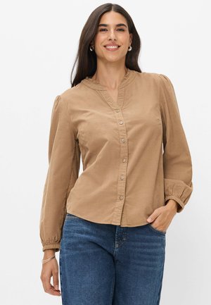 Lächelnde Frau mit langen dunklen Haaren, die eine beige geknöpfte Bluse mit gerafften Manschetten und blaue Jeans trägt, steht mit einer Hand in der Tasche.