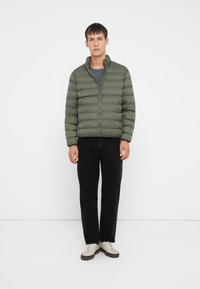 Grønn dunjakke med høy krage, glidelås og kviltet design. Båret over en grå skjorte, kombinert med svarte bukser og lyse joggesko.