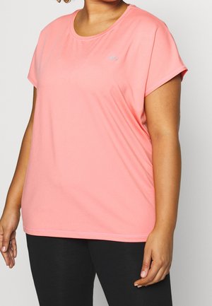 Osoba o krągłej sylwetce w krótkim koralowym sportowym T-shircie i czarnych legginsach, stojąca na tle jednolitego jasnego tła.