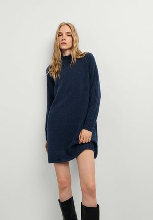 Junge Frau mit langen blonden Haaren, die ein marineblaues Strickkleid und schwarze kniehohe Stiefel trägt, steht vor einer schlichten hellen Wand.