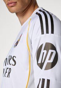 Maglia sportiva bianca con accenti neri e gialli, caratterizzata da tre strisce nere sulla spalla e un grande logo HP sulla manica.