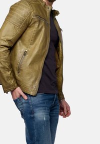 Giacca in pelle verde oliva con una texture liscia, chiusura con zip frontale e tasche, dettagli trapuntati sulle spalle, abbinata a una maglietta nera e jeans blu.