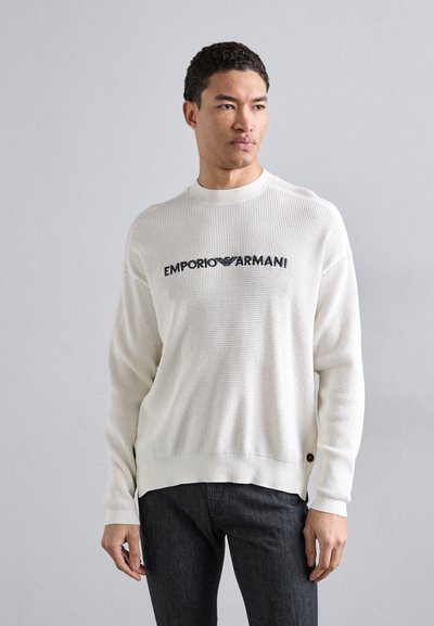 Sweatshirt en tricot texturé blanc arborant le logo "EMPORIO ARMANI" en noir, avec des poignets et un ourlet côtelés, des fentes sur les côtés et un col rond.