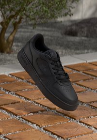 Svart lær sneaker med perforerte tær, rund form, flate lisser og Lacoste-logo på siden. Plassert på et mønstret flisebakgrunn.
