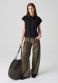 Camicetta nera a maniche corte con texture, pantaloni a gamba larga verde oliva e una grande borsa a rete nera. Le scarpe sono nere con un dettaglio a fiocco.