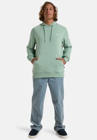 Mint green hoodie with a front kangaroo pocket, drawstring hood, ribbed cuffs, and a logo on the chest. Paired with light blue pants and dark shoes.