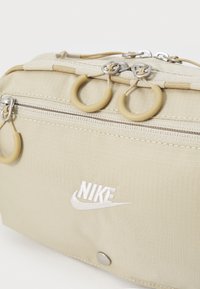 Nike-Tasche aus beigem Stoff mit Reißverschluss, gummierten Akzenten und besticktem Logo. Strukturierte Oberfläche mit verstärkten Hardware-Details.