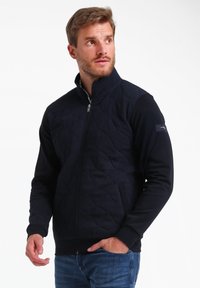 Veste zippée marine avec texture matelassée à l'avant, manches unies, col montant et écusson en tissu contrasté sur le bras gauche.