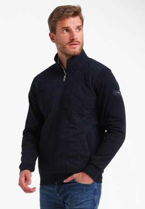 Marinefarbene Zip-Jacke mit gestepptem Vorderteil, einfarbigen Ärmeln, hohem Kragen und kontrastierendem gewebten Logo-Patch am linken Arm.