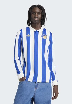 Giovane con i capelli intrecciati che indossa una maglia sportiva a maniche lunghe a righe blu e bianche e jeans blu, in piedi con una mano in tasca.